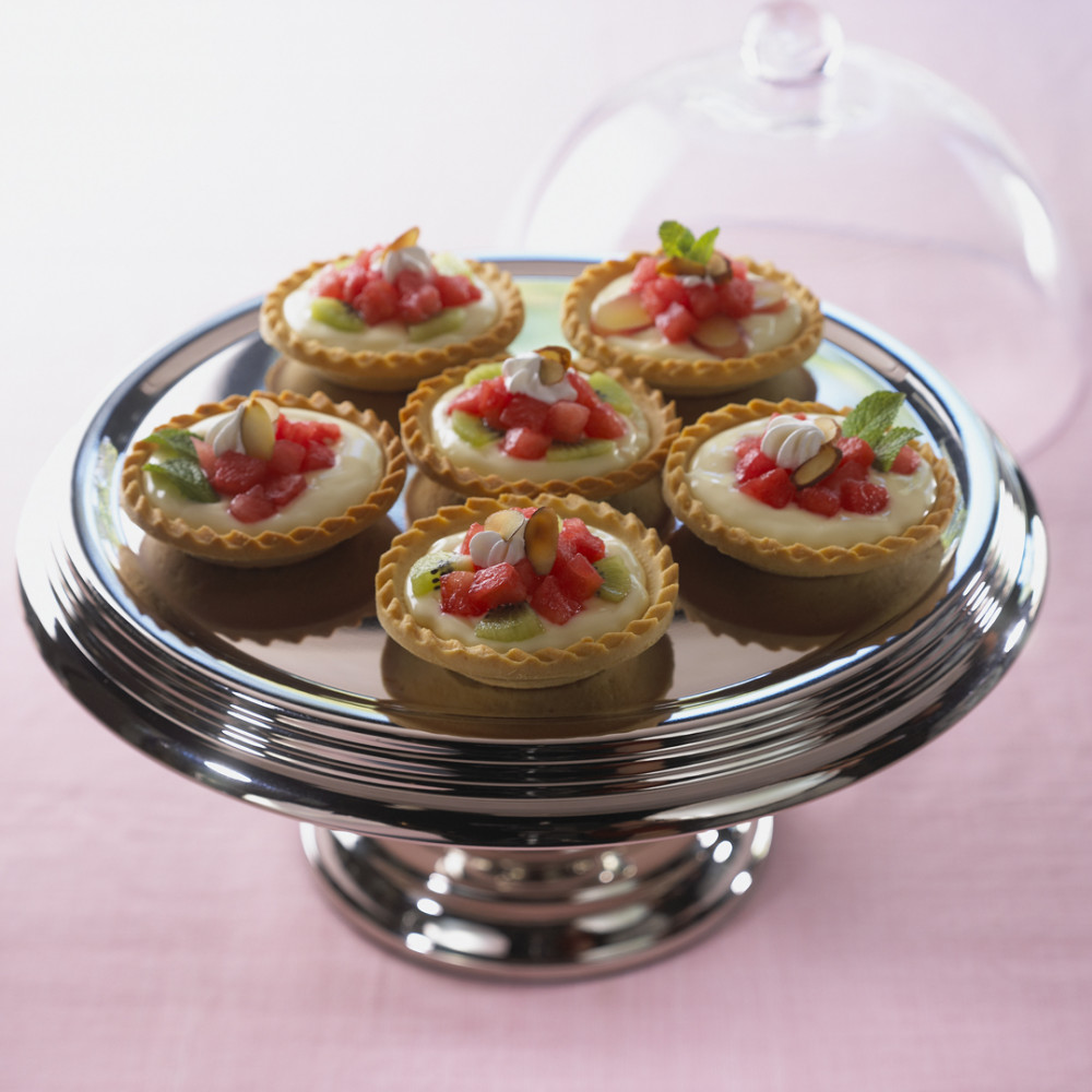 Six mini tarts on silver serving pedestal cake plate. Tarts garnished with slivered almonds, mint, diced watermelon and whipped cream.