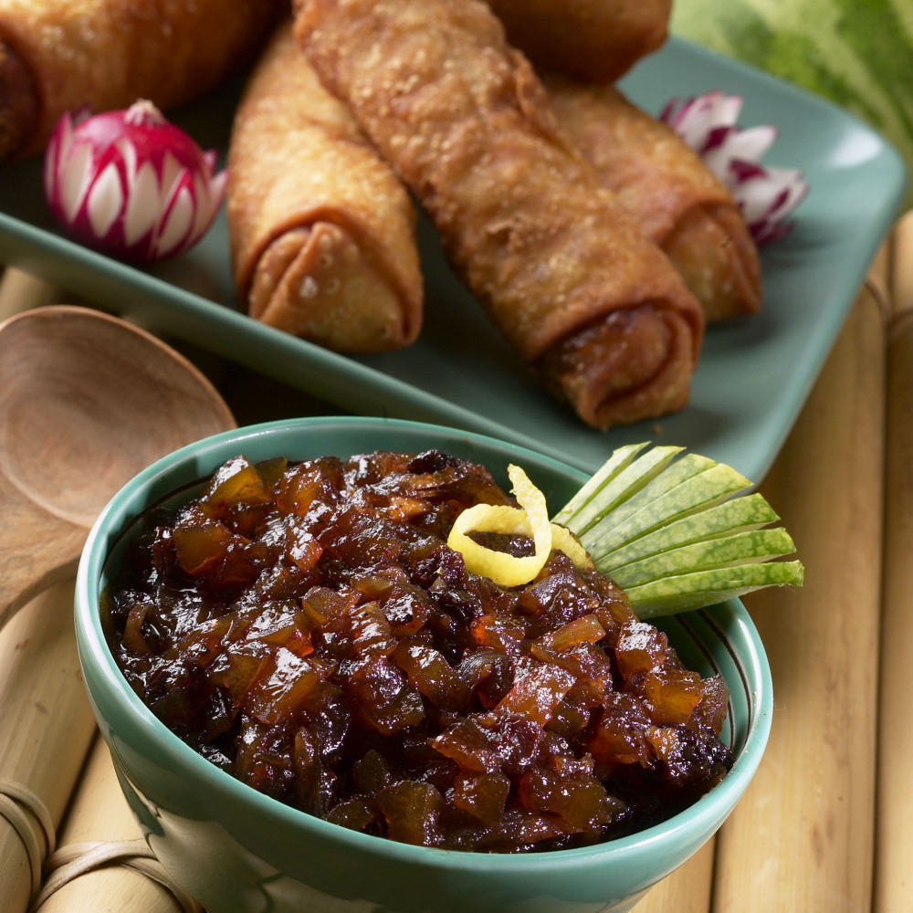 Walnut Currant Chutney in small bowl with decoratively cut watermelon rind and lemon zest as garnishes. Egg rolls and radishes on plate behind. Whole watermelon in background