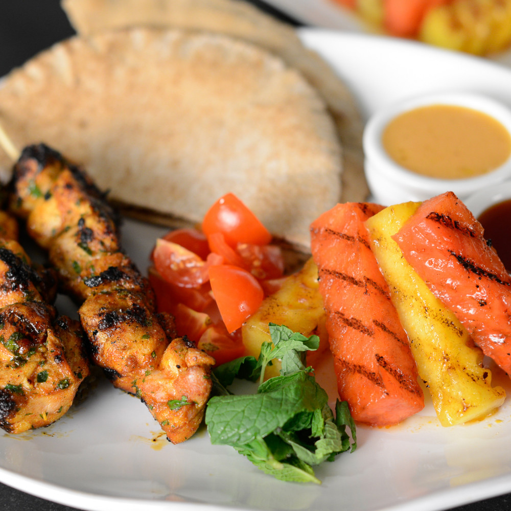 Watermelon Molasses Glazed Moroccan Chicken Kebabs set on plate with pita, cut tomatoes, grilled watermelon sticks and sauces.