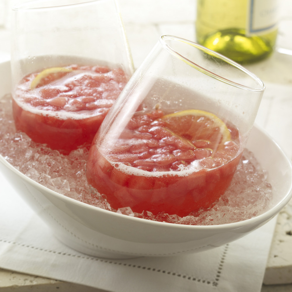 Watermelon Wine Spritzers set in oval dish sitting in crushed ice, garnished with lemon slice. Partial view of wine bottle in background.