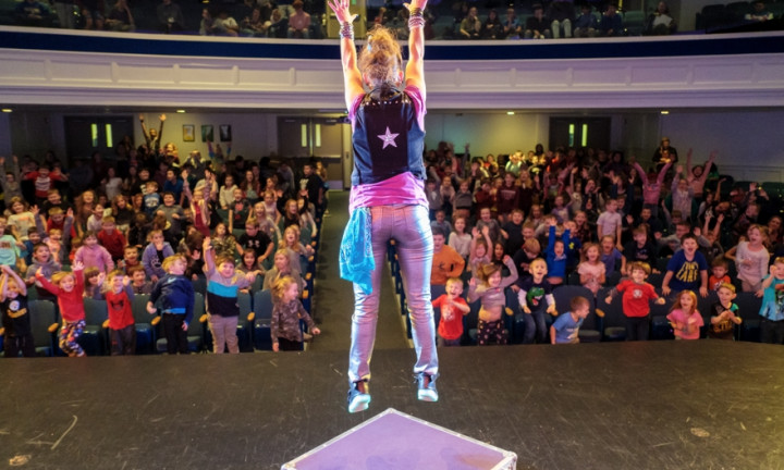 Jump with Jill, view from back of stage towards audience with performer center state, arms up