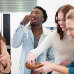 four people preparing a dish, one eating