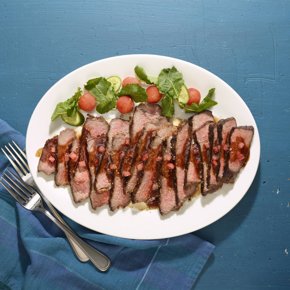 Flank steak with watermelon glaze on a serving plate with utensils.