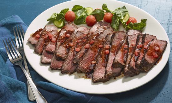 Flank steak with watermelon glaze on a serving plate with utensils.
