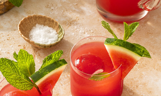 Two cups of watermelon juice with watermelon ice cubes garnished with watermelon slices. Small bowl of salt and pitcher with watermelon juice off to the side.