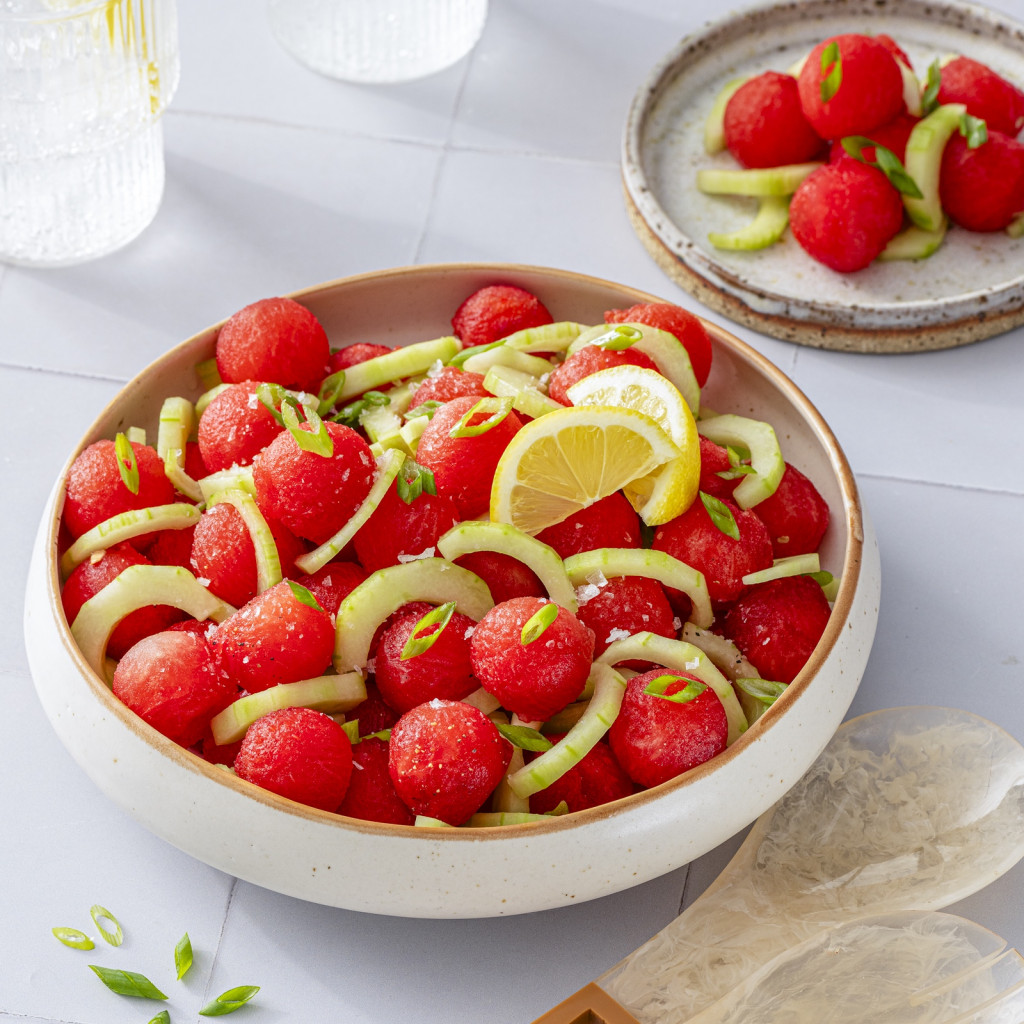 Watermelon balls with cucumber, scallions and lemon in a bowl with a serving size in the background.