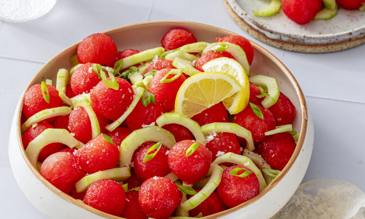 Watermelon balls with cucumber, scallions and lemon in a bowl with a serving size in the background.