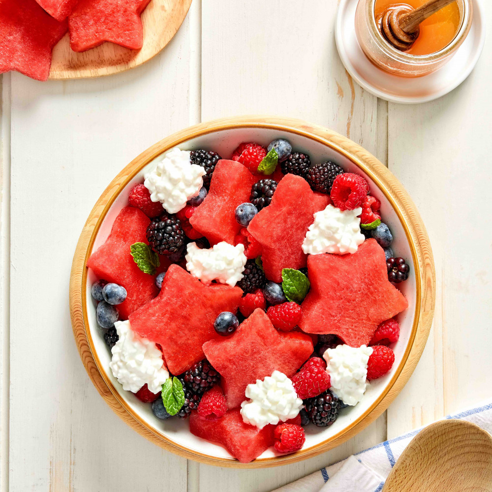 Fruit salad with star shaped watermelon cut outs, blackberries, blueberries, raspberries and cottage cheese with mint on top. Container of honey to drizzle on top.