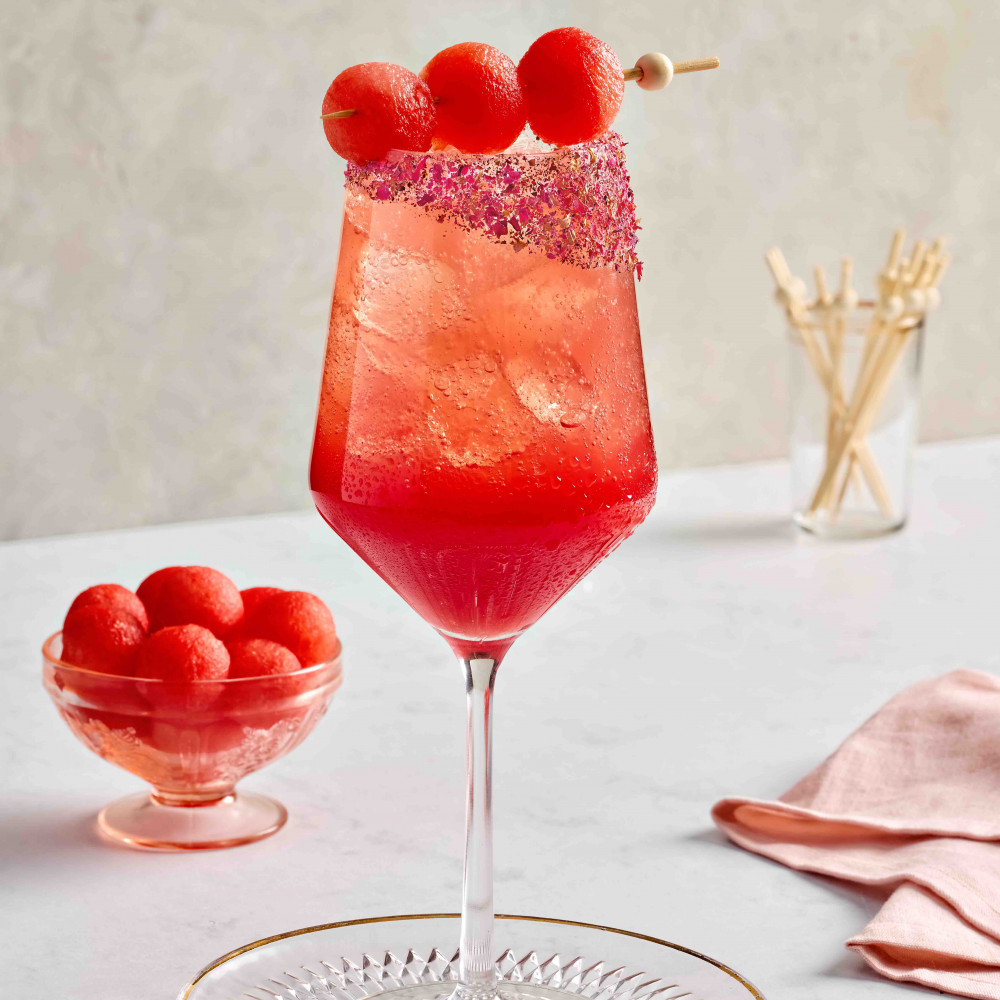 Watermelon spritz mocktail in a wine glass with crushed rose petals on the rim. Also has three melon balls on a skewer draped across the glass with melon balls in the background.