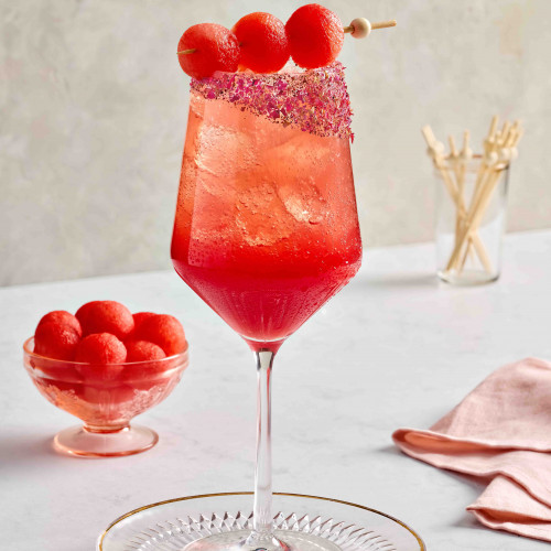 Watermelon spritz mocktail in a wine glass with crushed rose petals on the rim. Also has three melon balls on a skewer draped across the glass with melon balls in the background.