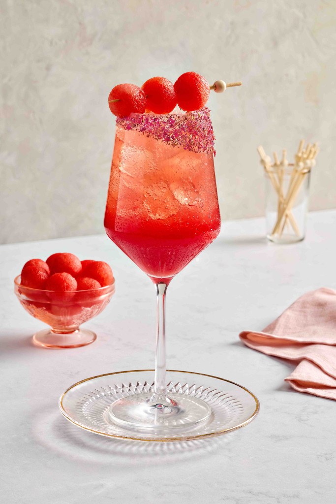 Watermelon spritz mocktail in a wine glass with crushed rose petals on the rim. Also has three melon balls on a skewer draped across the glass with melon balls in the background.