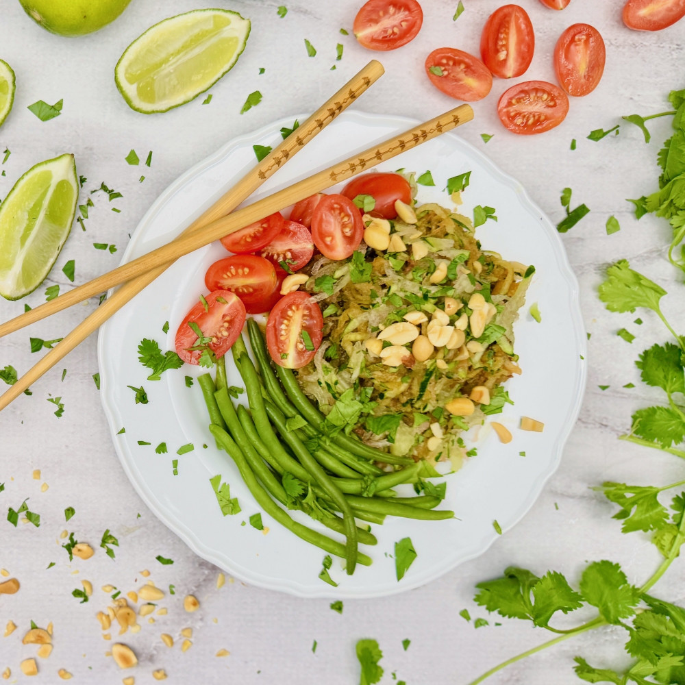 Watermelon rind salad inspired by a Thai Green papaya salad on white plate with green beans, rind slaw, tomatoes, peanuts and cilantro. Chopsticks are also on plate, lime wedges are off to the side.