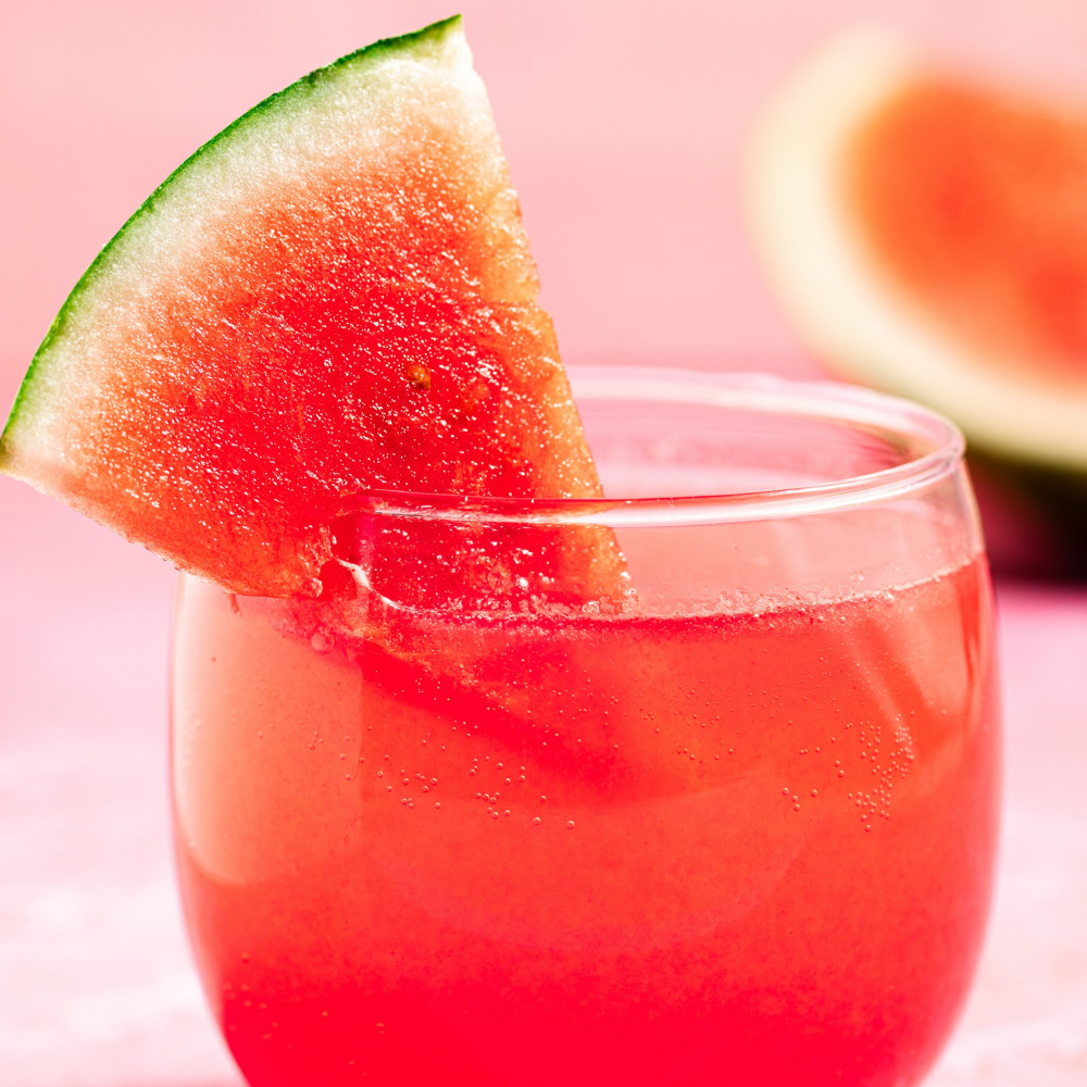 Red Watermelon Aperol Spritz in a clear, stemless glass with a small watermelon wedge on the rim