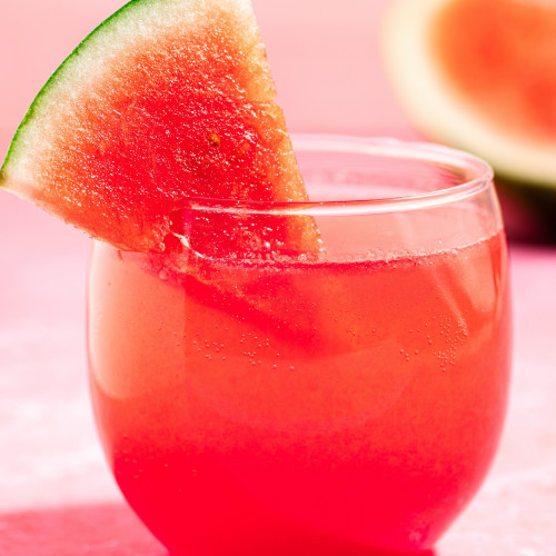 Red Watermelon Aperol Spritz in a clear, stemless glass with a small watermelon wedge on the rim