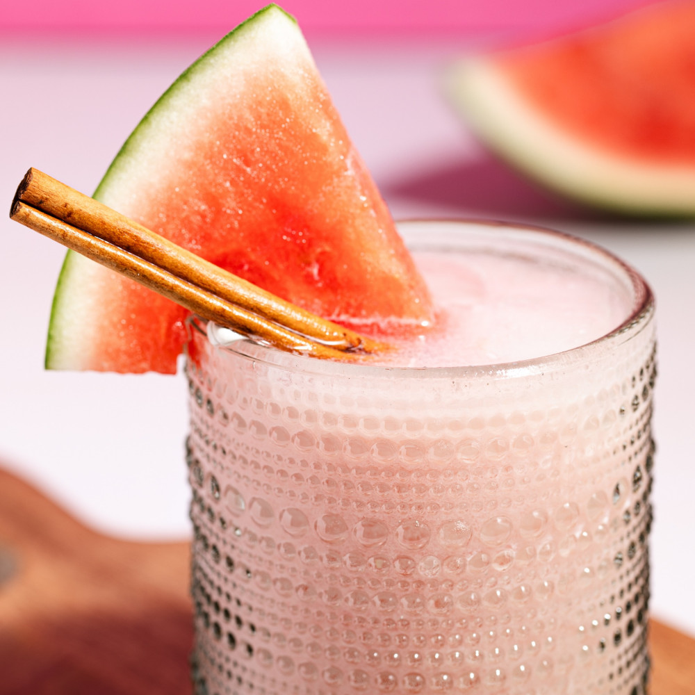 Pink margarita in a short pebbled glass on a wood serving board. Includes cinnamon stick and watermelon wedge garnish.