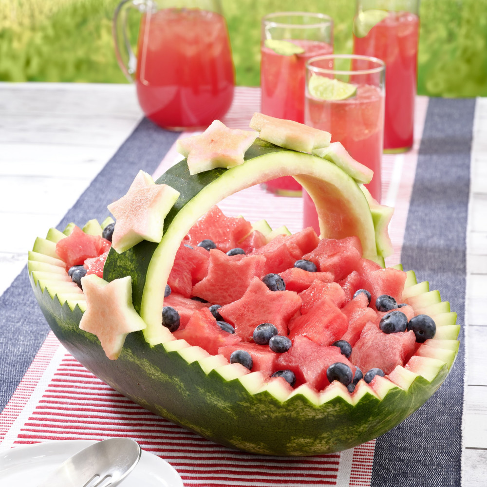 Watermelon carved into a basket filled with watermelon shaped stars and blueberries. Sitting on a blue and red runner next to a white plate with spoon and three glasses and pitcher of watermelon juice.