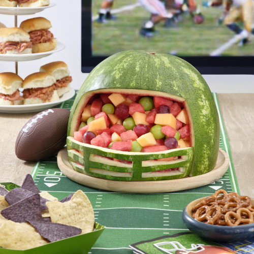 Watermelon carved into football helmet filled with fruit salad including watermelon, green and red grapes and cantaloupe in front of a TV and a 3-tiered tower with pork sliders.