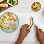 Two hands on a white cutting board cutting the green skin off of watermelon rind. Other cuts of watermelon rind are also on a blue plate on the cutting board, as well as a dish of lime wedges and watermelon cubes on a white plate.