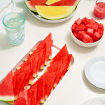 Red and yellow watermelon in a white bowl, watermelon cubes in a white bowl, rows of watermelon slices on a white vertical tray, two white circular plates and two glasses of water on a white tablescape.