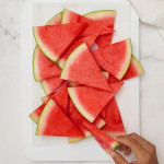 several watermelon wedge slices on a plate with white marble background. A hand is reaching in to grab a slice.