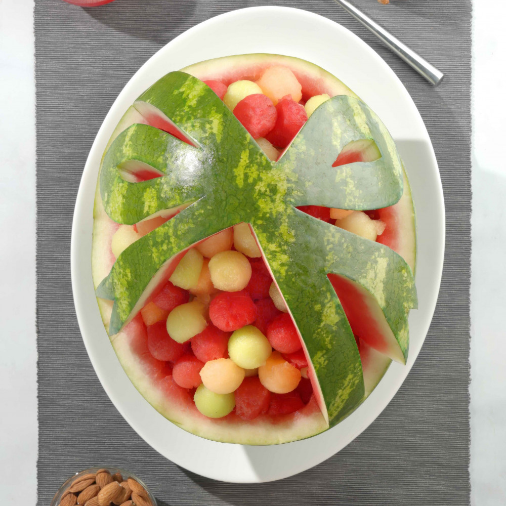 Ribbon Basket with watermelon balls inside basket/bowl on a white plate surrounded by two glasses of watermelon juice, a bowl of almonds and a bowl of pretzels.