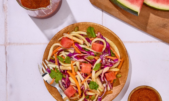 Rind Slaw with fresh watermelon cubes in a wooden bowl with fork featuring recipe elements around the dish including Tajin and lime.